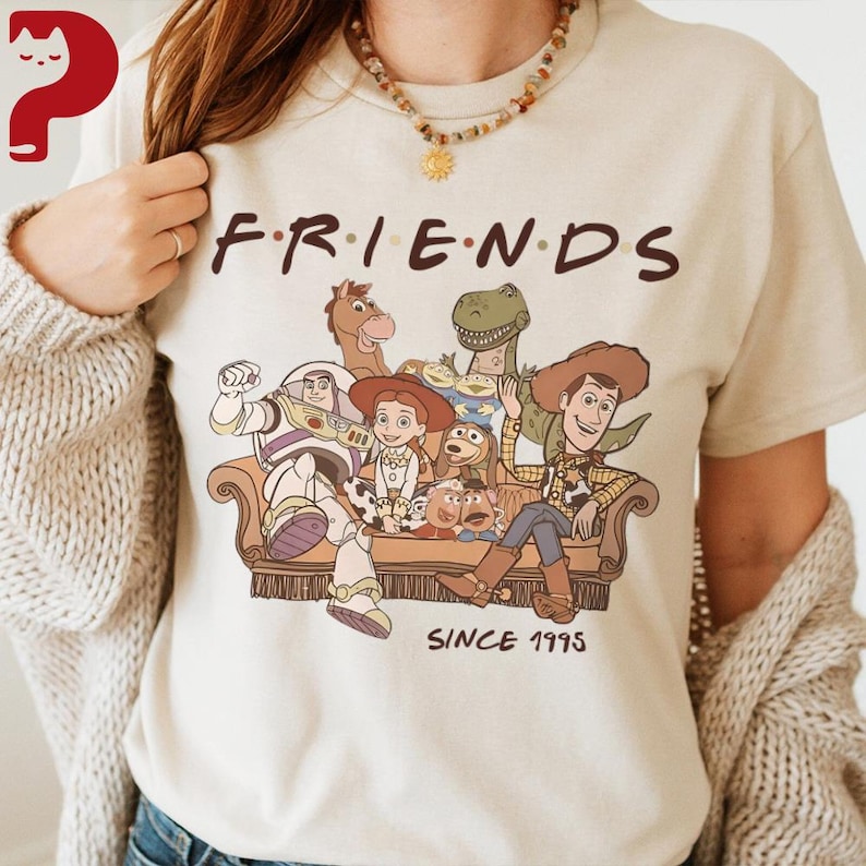 May include: Beige t-shirt featuring the word "FRIENDS" in brown letters above a graphic of Toy Story characters sitting on a couch. The text "SINCE 1995" is printed below the couch in brown letters.
