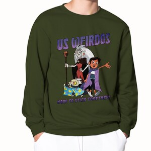 Op de afbeelding: Een groen sweatshirt met een grafische print van personages uit de animatieserie "The Owl House". De grafiek toont de personages Luz, Eda en King. De tekst op de grafiek luidt "Us Weirdos Have to Stick Together!"