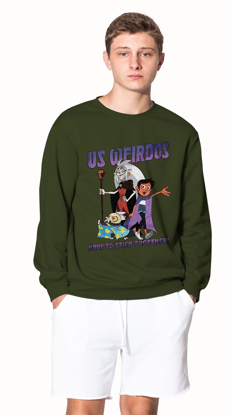 Op de afbeelding: Een groen sweatshirt met een grafische print van personages uit de animatieserie "The Owl House". De grafiek toont de personages Luz, Eda en King. De tekst op de grafiek luidt "Us Weirdos Have to Stick Together!"