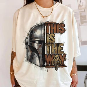 Może przedstawiać: Kremowa koszulka z nadrukiem w stylu vintage przedstawiającym hełm Mandaloriana i frazę "THIS IS THE WAY" w brązowych i złotych literach. Projekt zawiera efekty rozprysk&oacute;w farby.