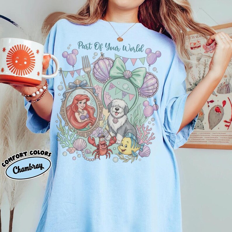 Może przedstawiać: Jasnoniebieska koszulka z kolorową grafiką przedstawiającą Ariel, psa i inne postacie z Małej Syrenki. Tekst "Part Of Your World" znajduje się nad grafiką. Koszulka jest oznaczona jako "Comfort Colors Chambray".