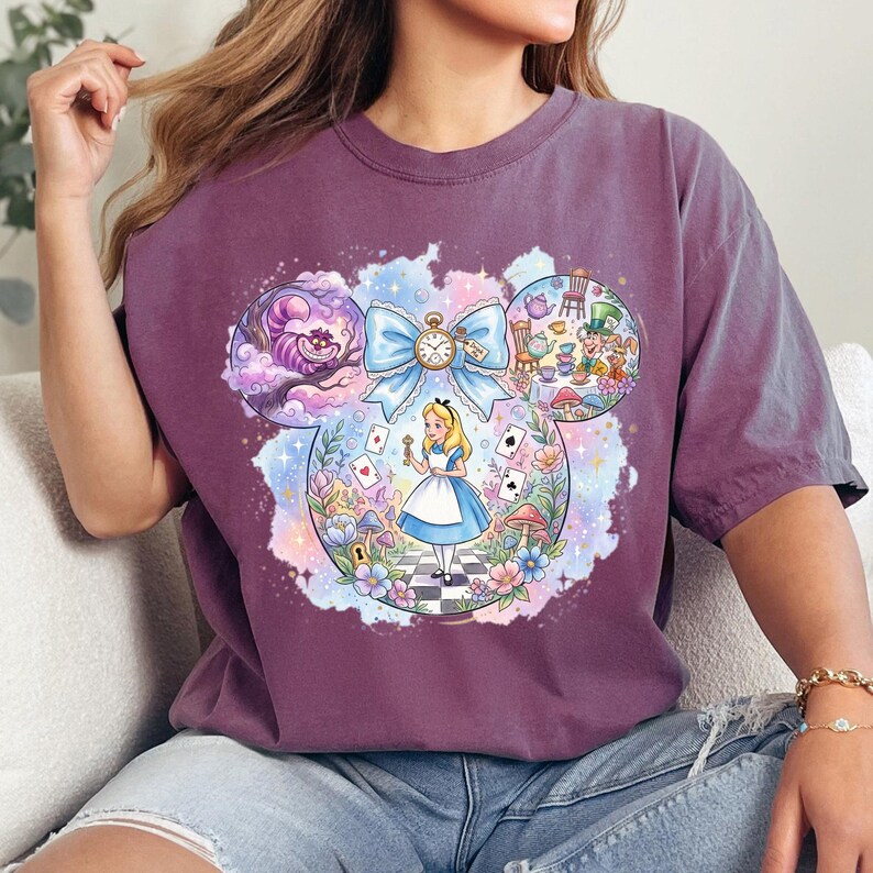 May include: A purple t-shirt with an Alice in Wonderland illustration. The design, set within a Mickey Mouse ear shape, features Alice, the Cheshire Cat, and other story elements. The artwork is colourful with a watercolour effect.