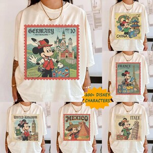 Op de afbeelding: Cr&egrave;mekleurige T-shirts met Mickey Mouse-ontwerpen met reisthema. Elk shirt toont een postzegel uit verschillende landen: Duitsland, China, Frankrijk, Verenigd Koninkrijk, Mexico en Itali&euml;. De shirts hebben de tekst "200+ DISNEY CHARACTERS".