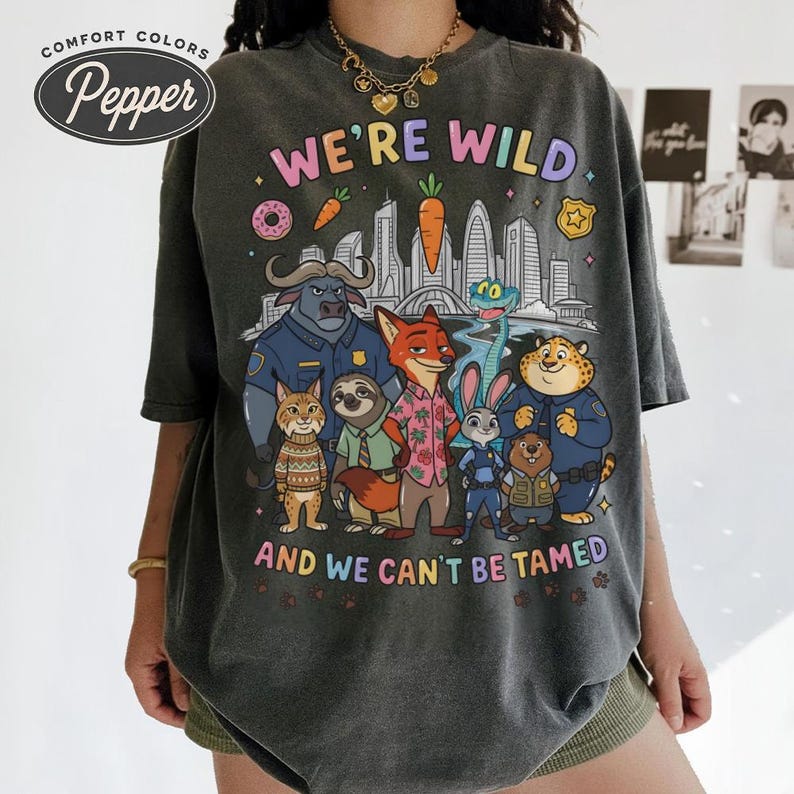 K&ouml;nnte beinhalten: Dunkelgraues Comfort Colors T-Shirt mit einer farbenfrohen Grafik mit Figuren aus einem beliebten Animationsfilm. Der Text "WE'RE WILD AND WE CAN'T BE TAMED" ist &uuml;ber den Figuren aufgedruckt. Das Shirt hat eine lockere Passform.