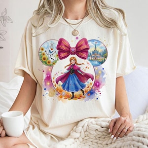Puede incluir: Camiseta color crema con un gr&aacute;fico colorido de una princesa con un vestido azul y una capa rosa, con un gran lazo rosa. El dise&ntilde;o incluye im&aacute;genes de un mu&ntilde;eco de nieve, girasoles y un castillo.