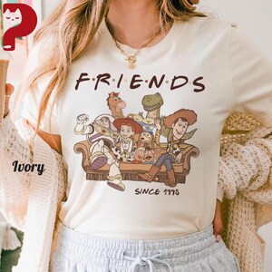 May include: Ivory-colored t-shirt featuring a graphic of Toy Story characters on a brown couch, with the word "FRIENDS" above and "SINCE 1995" below. The shirt is a crew neck.