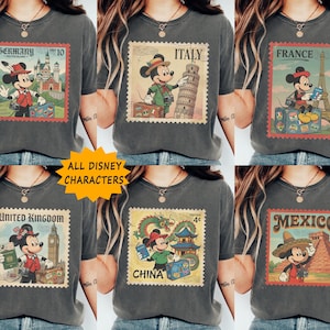 Op de afbeelding: Donkergrijze t-shirts met Mickey Mouse-reisontwerpen. Elk shirt toont een bezienswaardigheid van een ander land en Mickey Mouse in bijpassende kleding, gestyled als vintage postzegels. De tekst "ALL DISNEY CHARACTERS" is ook zichtbaar.