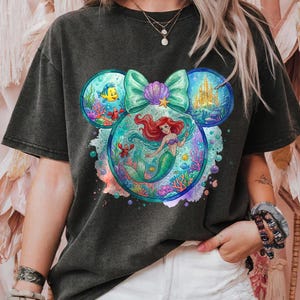 May include: Dark grey t-shirt with a colourful graphic of Ariel the mermaid within a Mickey Mouse head shape. The design includes a teal bow, a castle, and sea creatures. The shirt has short sleeves.