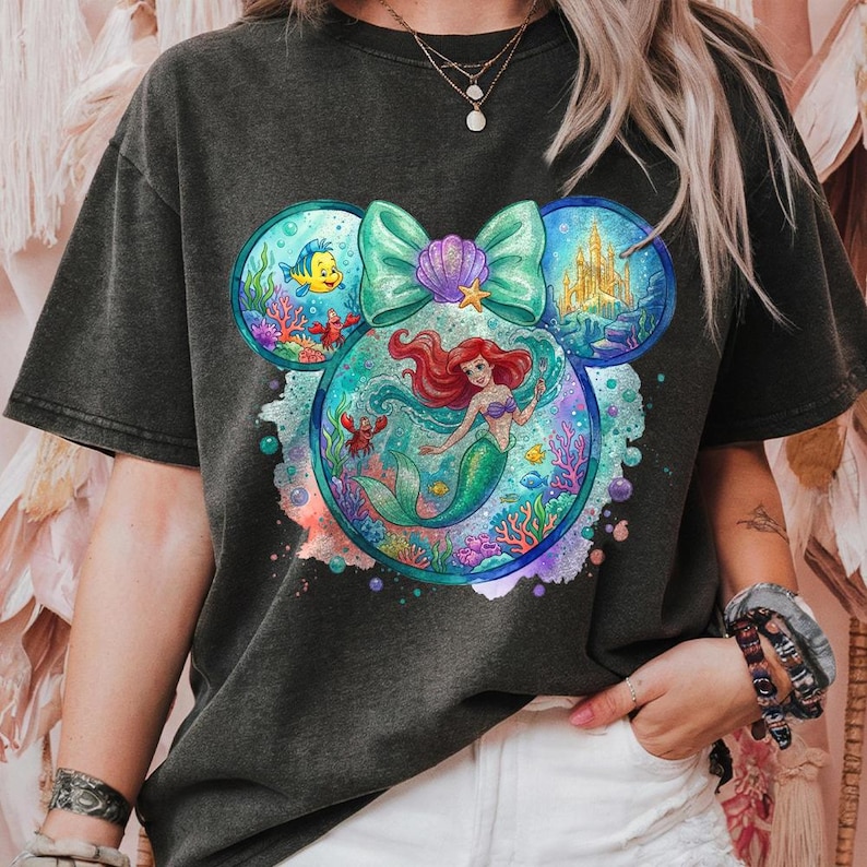 May include: Dark grey t-shirt with a colourful graphic of Ariel the mermaid within a Mickey Mouse head shape. The design includes a teal bow, a castle, and sea creatures. The shirt has short sleeves.