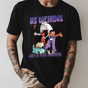 Op de afbeelding: Zwart T-shirt met een grafische print van cartoonfiguren uit de show "The Owl House". De tekst "US WEIRDOS" is in paars boven de figuren gedrukt. De tekst "HAVE TO STICK TOGETHER!" is onder de figuren gedrukt.