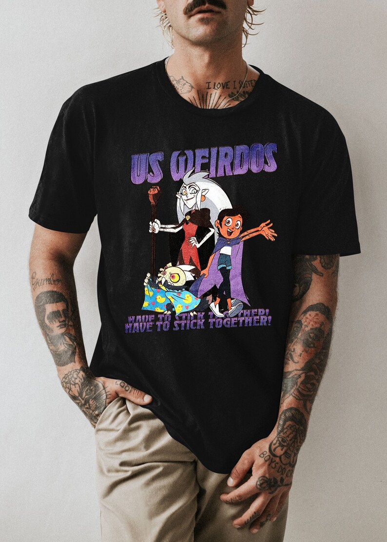 Op de afbeelding: Zwart T-shirt met een grafische print van cartoonfiguren uit de show "The Owl House". De tekst "US WEIRDOS" is in paars boven de figuren gedrukt. De tekst "HAVE TO STICK TOGETHER!" is onder de figuren gedrukt.