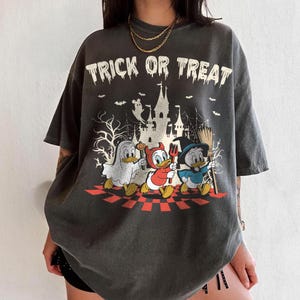 May include: Dark gray t-shirt with a Halloween graphic. The shirt features the text "TRICK OR TREAT" above a cartoon image of three ducks in costumes walking in front of a haunted castle.
