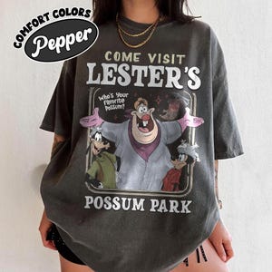May include: Dark gray t-shirt with a graphic featuring the text "COME VISIT LESTER'S POSSUM PARK". The graphic includes cartoon characters and the phrase "Who's Your Favorite Possum?" The shirt has short sleeves and a crew neck.