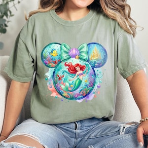 May include: Sage green t-shirt with a colourful graphic of Ariel from The Little Mermaid. The design is in the shape of Mickey Mouse ears, with Ariel in the centre, surrounded by sea creatures and a castle. A light green bow and seashell complete the design.