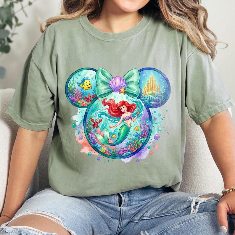 May include: Sage green t-shirt with a colourful graphic of Ariel from The Little Mermaid. The design is in the shape of Mickey Mouse ears, with Ariel in the centre, surrounded by sea creatures and a castle. A light green bow and seashell complete the design.