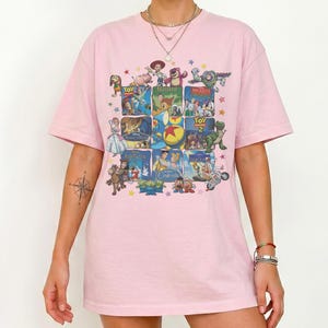 May include: A light pink t-shirt featuring a collage of animated movie posters. The design includes images from classic films like "Toy Story," "Bambi," and "Cinderella." The shirt has a crew neck and short sleeves.