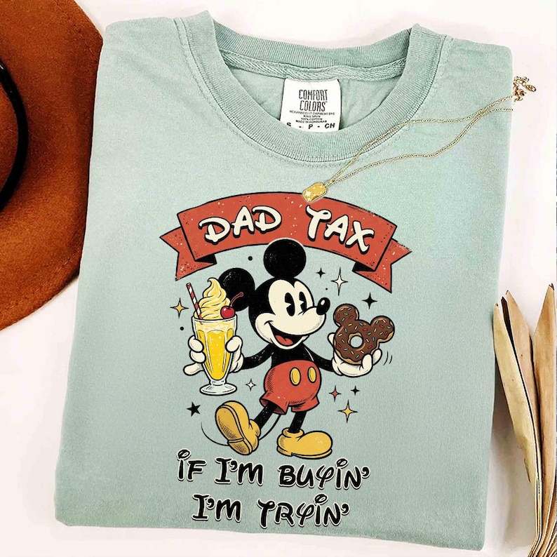 Puede incluir: Camiseta Comfort Colors azul claro con un gr&aacute;fico de Mickey Mouse sosteniendo un batido y una rosquilla. El texto "DAD TAX" est&aacute; encima de Mickey, y "If I'm buyin' I'm tryin'" est&aacute; debajo. Un collar de oro est&aacute; sobre la camiseta.
