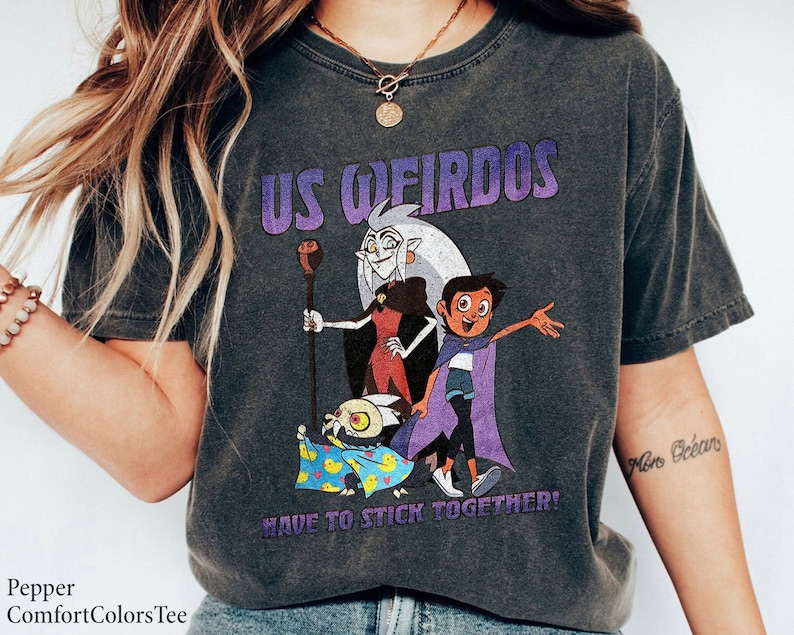 Op de afbeelding: Een donkergrijs T-shirt met een grafische print van personages uit de animatieserie "The Owl House". De grafiek toont de personages Luz, Eda en King. De tekst "Us Weirdos Have to Stick Together!" is onder de grafiek gedrukt. Het shirt heeft een verweerde look.