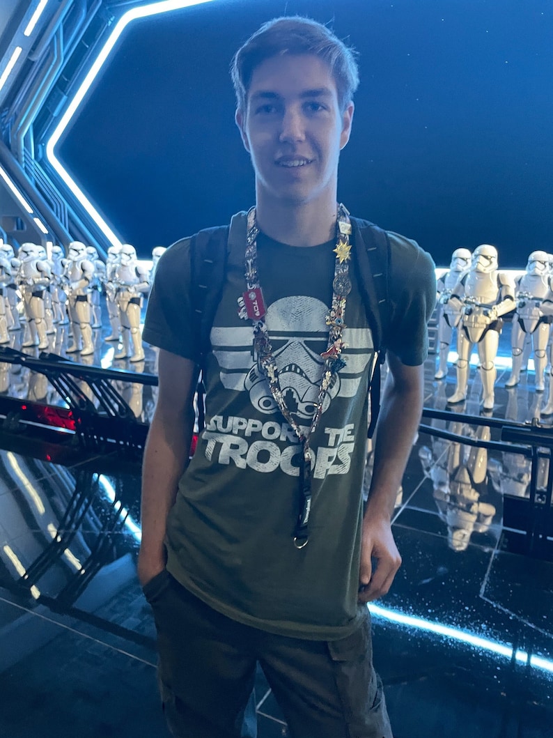May include: A young person wearing a green t-shirt with the text "Support the Troops" and a white stormtrooper graphic. They are wearing a lanyard with multiple pins and charms. The person is standing in front of a backdrop of white stormtroopers.