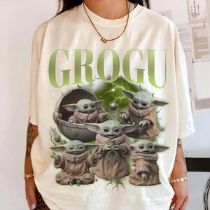 May include: Cream-colored t-shirt featuring the word "GROGU" in green, with a graphic of Grogu, also known as Baby Yoda, in various poses. The design includes Grogu in a pod and holding a frog. The shirt has a relaxed fit.