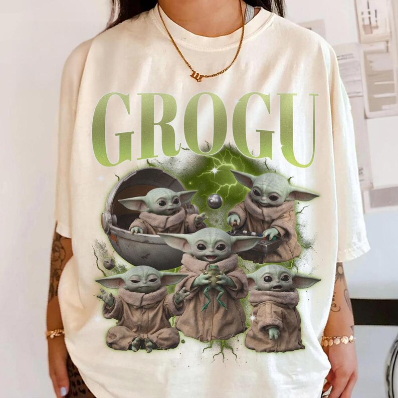 May include: Cream-colored t-shirt featuring the word "GROGU" in green, with a graphic of Grogu, also known as Baby Yoda, in various poses. The design includes Grogu in a pod and holding a frog. The shirt has a relaxed fit.