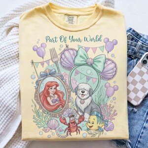 Może przedstawiać: Jasnoż&oacute;łta koszulka z kolorową grafiką i napisem "Part Of Your World". Projekt zawiera Ariel, psa i inne postacie. Koszulka ma okrągły dekolt i kr&oacute;tkie rękawy.