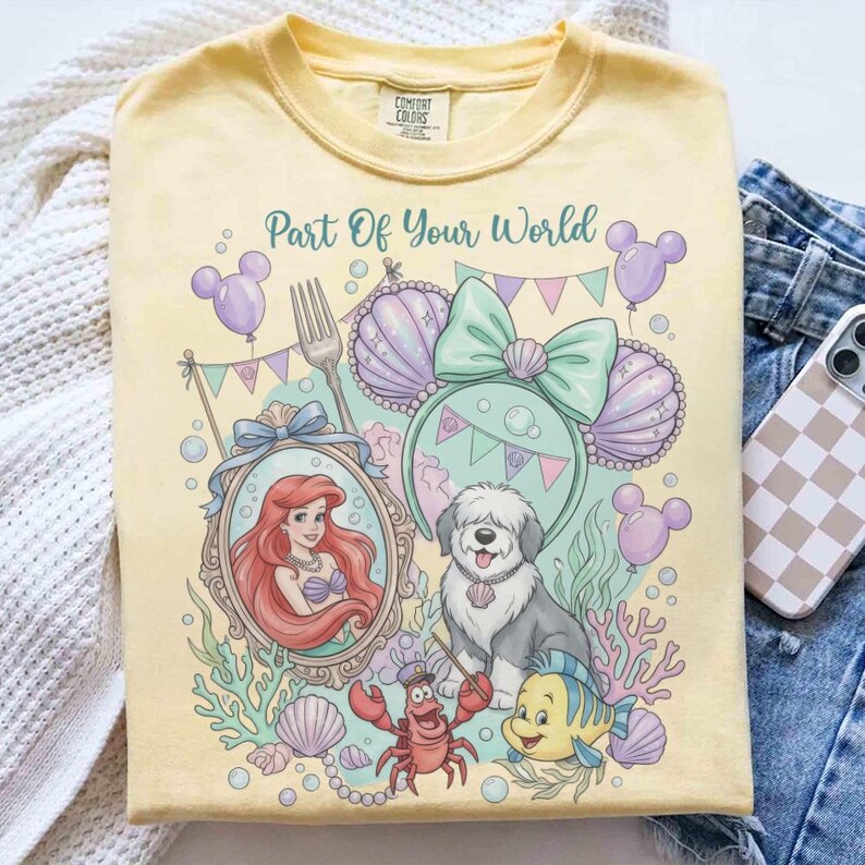 Może przedstawiać: Jasnoż&oacute;łta koszulka z kolorową grafiką i napisem "Part Of Your World". Projekt zawiera Ariel, psa i inne postacie. Koszulka ma okrągły dekolt i kr&oacute;tkie rękawy.