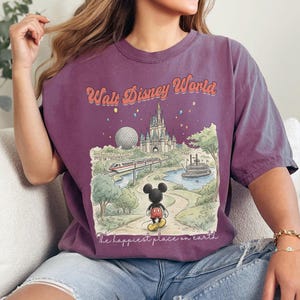 May include: A purple t-shirt featuring a vintage-style illustration of Walt Disney World. The design includes the park's castle, Spaceship Earth, a monorail, and Mickey Mouse. Text reads "Walt Disney World" and "The happiest place on earth."