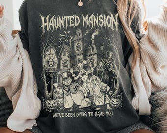 May include: Dark gray sweatshirt featuring a Haunted Mansion graphic. The design includes a spooky house, characters, and the text "Haunted Mansion" and "We've been dying to have you." The sweatshirt has a vintage, distressed look.