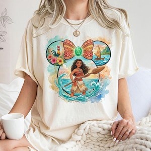 以下が含まれることがあります： クリーム色のTシャツ。海波の中でパドルを持っている漫画のキャラクターのカラフルなグラフィックが特徴です。デザインには、リボン、雄鶏、花、ヨットが含まれています。シャツは半袖です。