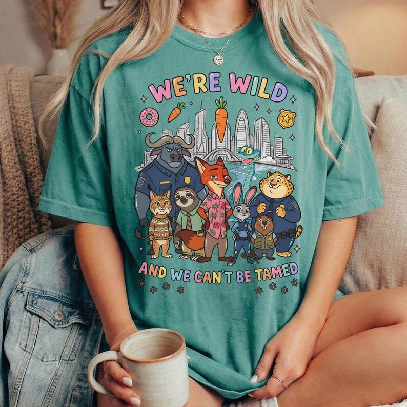 K&ouml;nnte beinhalten: T&uuml;rkisfarbenes T-Shirt mit dem Text "WE'RE WILD AND WE CAN'T BE TAMED" und einer Grafik von Zeichentricktieren und einer Skyline. Das Shirt hat einen Rundhalsausschnitt und kurze &Auml;rmel.