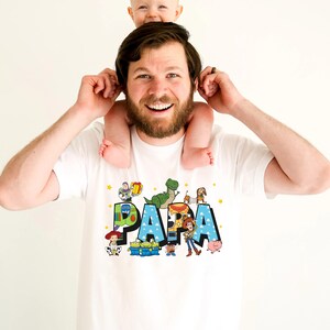 Puede incluir: Camiseta blanca con un gr&aacute;fico azul, verde y amarillo de la palabra "PAPA" con personajes de la franquicia de pel&iacute;culas Toy Story.