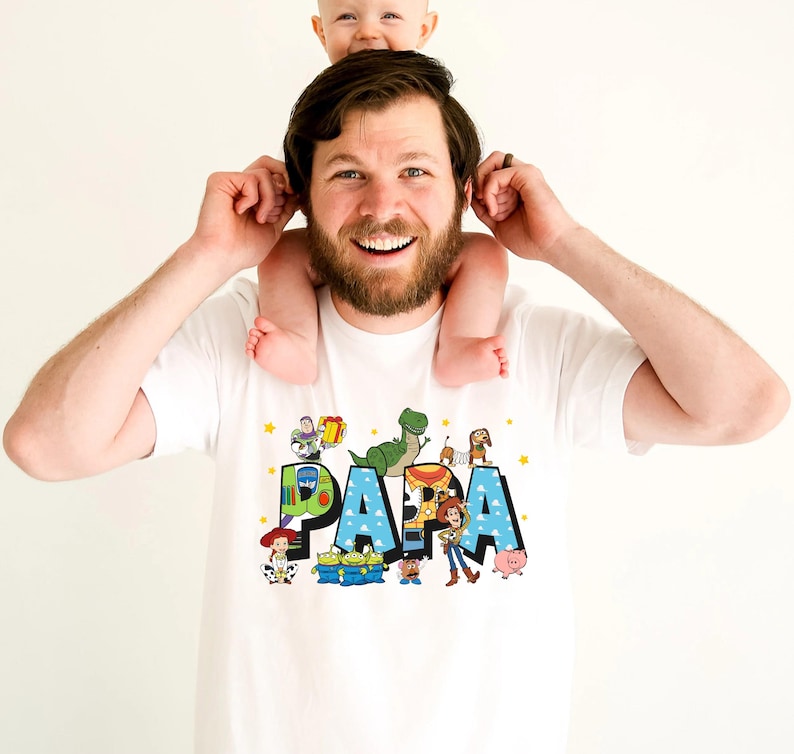 Puede incluir: Camiseta blanca con un gr&aacute;fico azul, verde y amarillo de la palabra "PAPA" con personajes de la franquicia de pel&iacute;culas Toy Story.