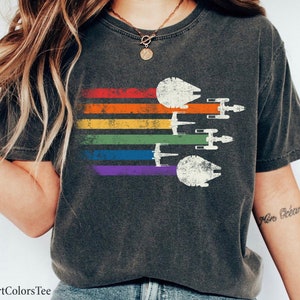 May include: A dark grey t-shirt featuring a rainbow stripe design and a vintage-style image of the Millennium Falcon and X-Wing starfighters from Star Wars. The text "More Ocean" is written on the right sleeve.