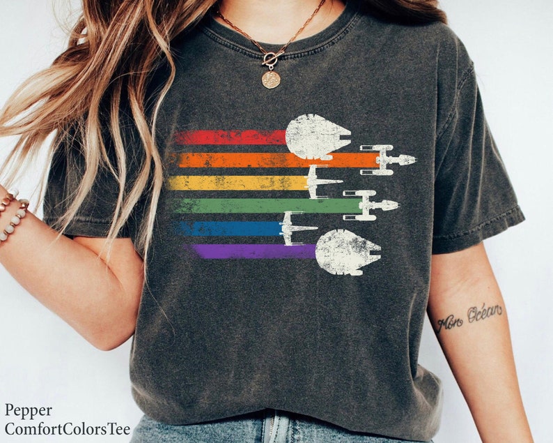 May include: A dark grey t-shirt featuring a rainbow stripe design and a vintage-style image of the Millennium Falcon and X-Wing starfighters from Star Wars. The text "More Ocean" is written on the right sleeve.