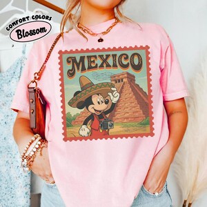 Op de afbeelding: Roze T-shirt met een afbeelding van Mickey Mouse met een sombrero, die een camera vasthoudt voor een piramide, met het woord "MEXICO" in een postzegelontwerp. Het shirt heeft ook de woorden "COMFORT COLORS" en "Blossom".