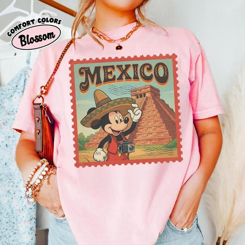 Op de afbeelding: Roze T-shirt met een afbeelding van Mickey Mouse met een sombrero, die een camera vasthoudt voor een piramide, met het woord "MEXICO" in een postzegelontwerp. Het shirt heeft ook de woorden "COMFORT COLORS" en "Blossom".