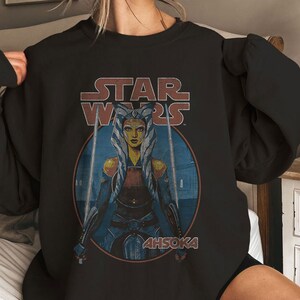 Op de afbeelding: Zwart sweatshirt met een Star Wars-afbeelding met Ahsoka Tano, een Togruta Jedi, die twee lichtzwaarden vasthoudt. De tekst "STAR WARS" staat boven haar en "AHSOKA" eronder.