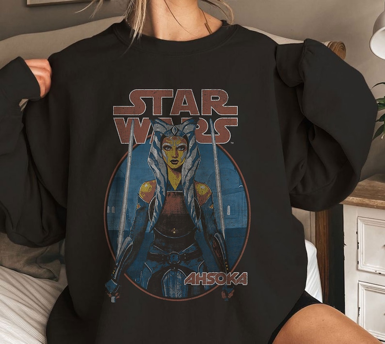Op de afbeelding: Zwart sweatshirt met een Star Wars-afbeelding met Ahsoka Tano, een Togruta Jedi, die twee lichtzwaarden vasthoudt. De tekst "STAR WARS" staat boven haar en "AHSOKA" eronder.