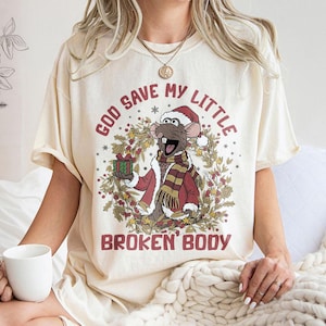 May include: Cream-colored t-shirt featuring a cartoon rat in a Santa hat and scarf, holding a gift. The text "GOD SAVE MY LITTLE BROKEN BODY" is printed above and below the rat illustration. The shirt has short sleeves.