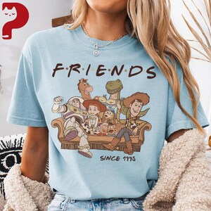 May include: Light blue t-shirt featuring the word "FRIENDS" in a stylized font, with a graphic of characters from the Toy Story movie. The graphic includes Woody, Buzz Lightyear, and other characters. The text "SINCE 1995" is below the graphic.