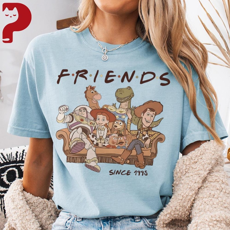 May include: Light blue t-shirt featuring the word "FRIENDS" in a stylized font, with a graphic of characters from the Toy Story movie. The graphic includes Woody, Buzz Lightyear, and other characters. The text "SINCE 1995" is below the graphic.