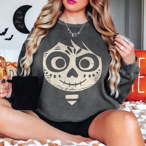 May include: A dark gray sweatshirt with a white graphic of a character from the movie Coco. The character has a skull face with a smile and large eyes. The sweatshirt has a relaxed fit and long sleeves.