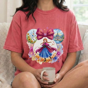 Puede incluir: Camiseta roja con un dise&ntilde;o gr&aacute;fico colorido de una princesa de Disney, un gran lazo y im&aacute;genes de un mu&ntilde;eco de nieve, un castillo y hojas de oto&ntilde;o. El dise&ntilde;o est&aacute; dentro de la forma de una oreja de Mickey Mouse.