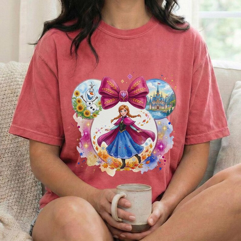 Puede incluir: Camiseta roja con un dise&ntilde;o gr&aacute;fico colorido de una princesa de Disney, un gran lazo y im&aacute;genes de un mu&ntilde;eco de nieve, un castillo y hojas de oto&ntilde;o. El dise&ntilde;o est&aacute; dentro de la forma de una oreja de Mickey Mouse.