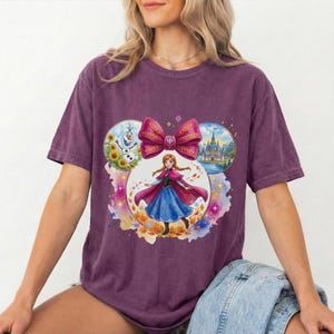 Puede incluir: Una camiseta morada con un gr&aacute;fico de una princesa con un vestido azul y una capa rosa, rodeada por un lazo e im&aacute;genes circulares de un mu&ntilde;eco de nieve, un castillo y flores. La camiseta tiene cuello redondo y mangas cortas.