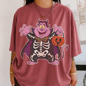 May include: A dusty rose short-sleeve t-shirt featuring a graphic print of a purple cartoon character wearing a black skeleton costume with a cape and holding an orange jack-o'-lantern bucket with a black handle and a tail.
