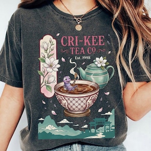 May include: Dark gray t-shirt featuring a whimsical design for "Cri-Kee Tea Co." with a cricket in a teacup, a teapot, and floral accents. The text "Est. 1998" is also included in the design.