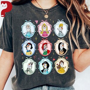 May include: Dark gray t-shirt featuring a grid of cartoon characters in oval frames with bows. The characters include a snowman, princesses, and other figures. The frames are in various pastel colors, with hearts and stars scattered around the design.