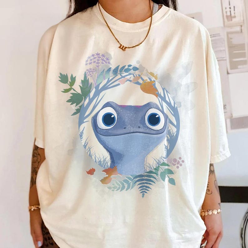 Puede incluir: Camiseta color crema con un dise&ntilde;o de rana azul de dibujos animados, rodeada por una corona floral. La cara de la rana es el punto central, con detalles en azul, verde y naranja. La camiseta es de corte holgado.